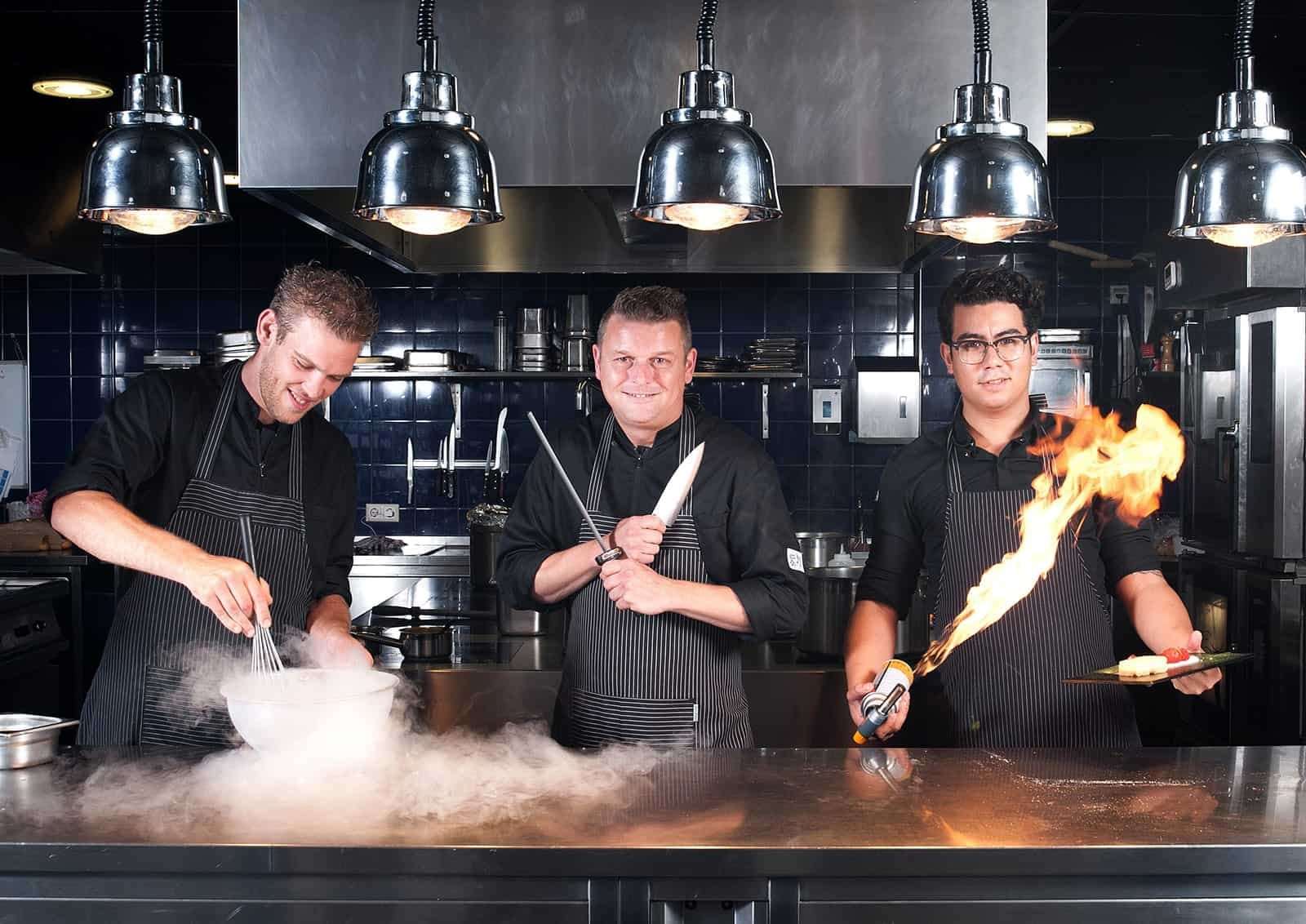 Drie koks aan het werk in de open keuken, een van hen flambeert